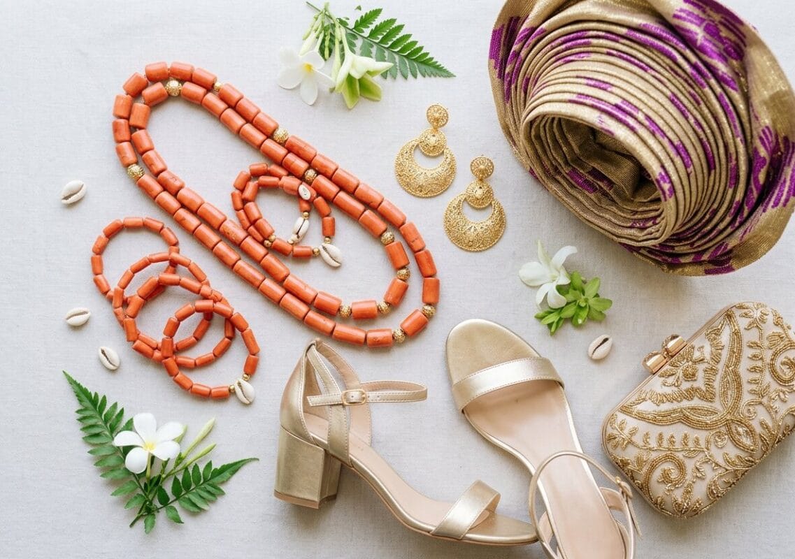 Nigerian wedding guest accessories including gold earrings, coral beads, clutch bag, and gele headwrap